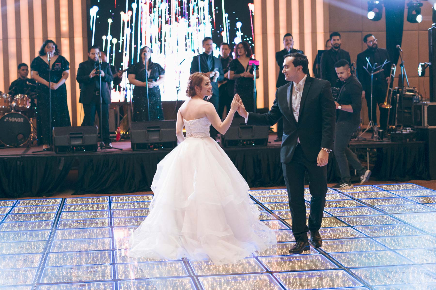 Novia entrando a la iglesia acompañada por sus padres; lleva velo y ramo de flores blancas, rodeada de invitados.