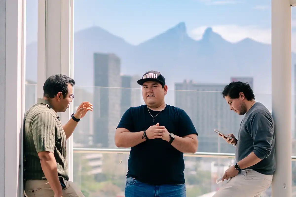 Tres hombres conversando relajadamente en una terraza con vista a Monterrey y el Cerro de la Silla al fondo.