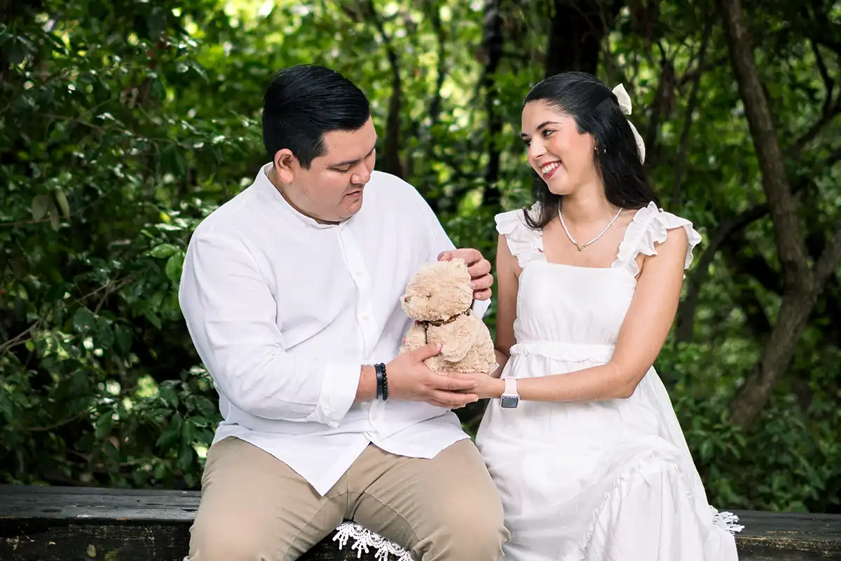 Pareja embarazada sentada en el bosque, sosteniendo un osito de peluche con una expresión tierna.