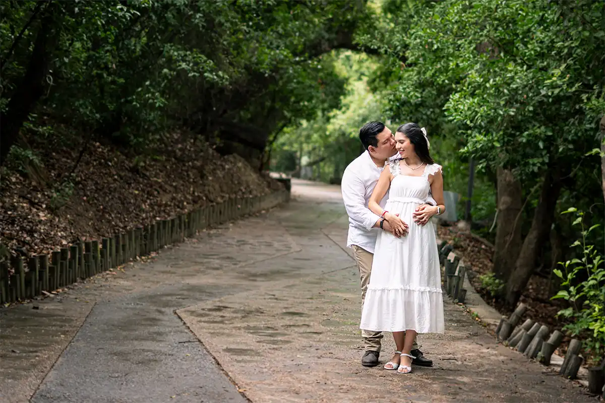 Pareja abrazada en un camino rodeado de árboles, disfrutando de un momento romántico en su sesión de embarazo.